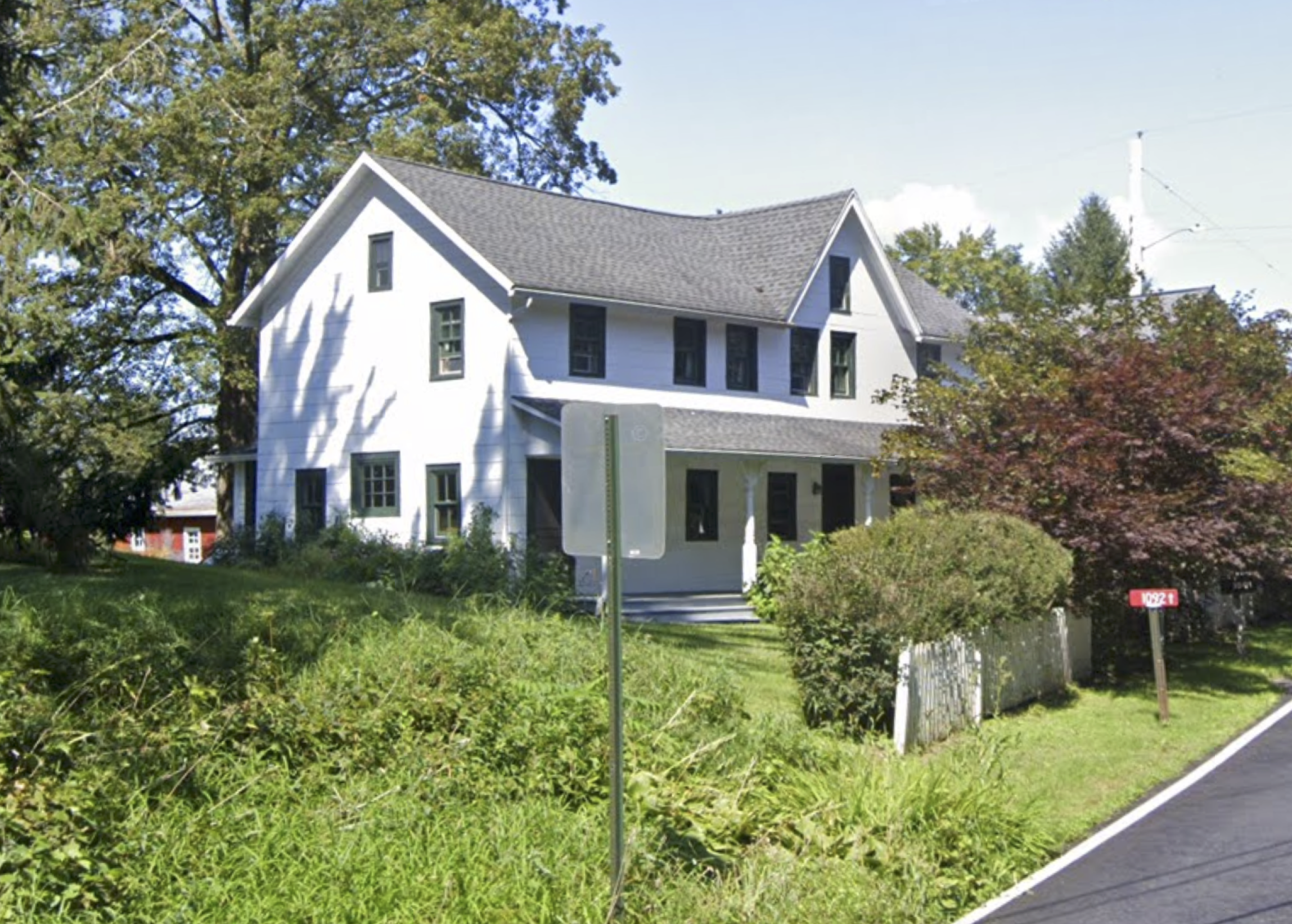 A white farm house partially obstructed by shrubbery growing along the fence.